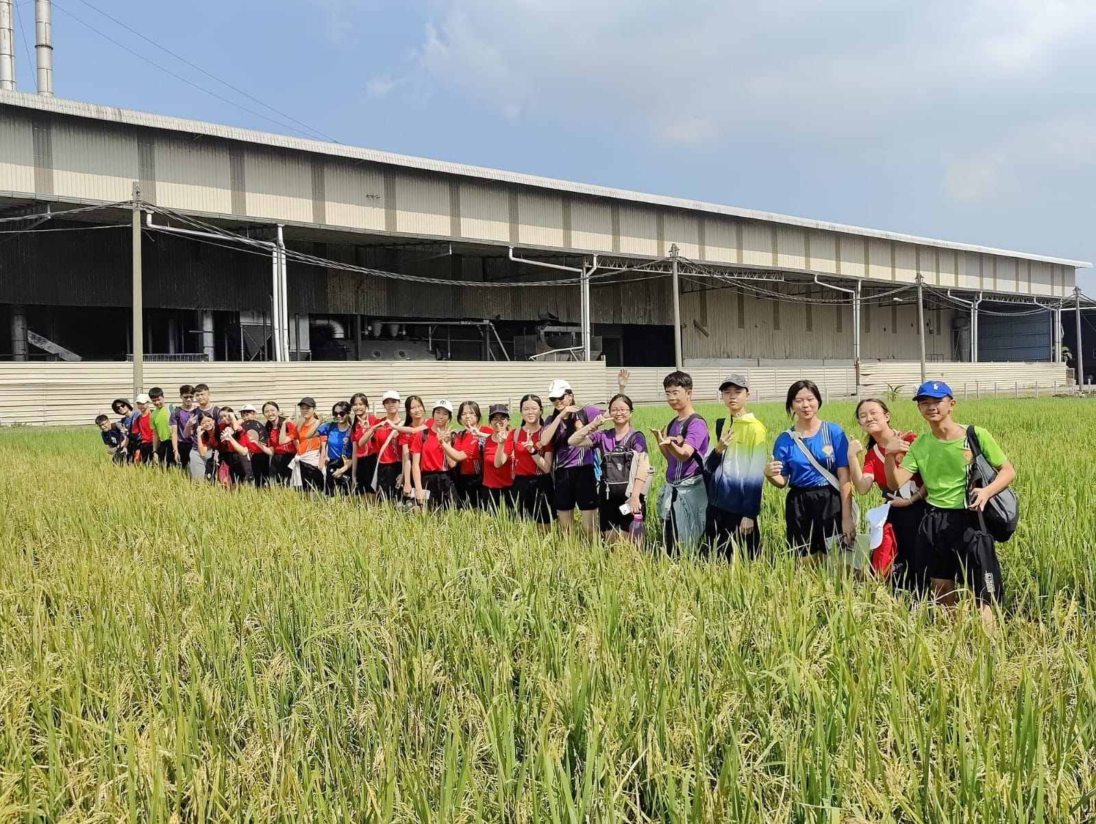 A group of students posing and smiling in a green rice field in front of a large industrial building.
